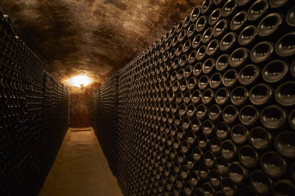 Wine cellar tunnel lined with hundreds of dark bottles stored in wooden racks, illuminated by a warm light at the end