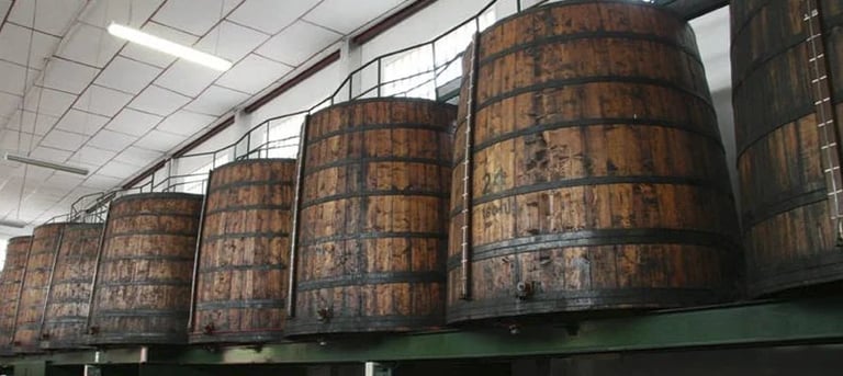 Large wooden barrels lined up on green metal support beams in an industrial warehouse setting with fluorescent ceiling lights